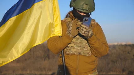 Tuinposter Female ukrainian army soldier using smartphone to read news or messaging with family. Girl in military uniform and helmet holding waving flag of Ukraine. Victory against russian aggression. Close up © olehslepchenko