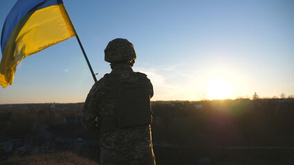 Soldier of ukrainian army stands at hill and holds waving flag of Ukraine. Man in military uniform...