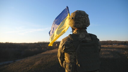 Ukrainian army soldier holding waving flag of Ukraine. Portrait of man in military uniform and helmet lifted up flag in hill. Victory against russian aggression. Invasion resistance concept. Slow mo - obrazy, fototapety, plakaty