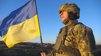 Ukrainian army soldier holding waving flag of Ukraine. Portrait of man in military uniform and helmet lifted up flag in hill. Victory against russian aggression. Invasion resistance concept. Slow mo