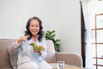 Obraz na płótnie Canvas Asian senior woman sitting eating vegetable salad and healthy food and eating happily on the sofa in the house for a healthy body. healthy food concept