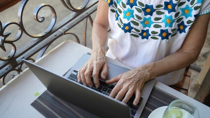 Closeup looking down on a mature old womans hands using a laptop computer with a drink on a table. Concept of digital nomad traveling the world.