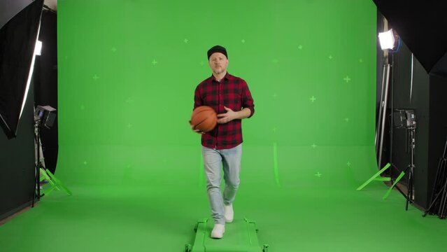 Portrait of Young Motivated Bearded Man in blue jeans, a black baseball cap and a plaid shirt, carries a basketball in his hands and walks forward against the background of a Green Screen, Chroma Key