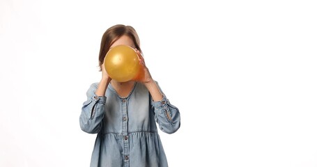 Pretty little girl in casual denim dress blowing, inflate yellow balloon on white background, studio
