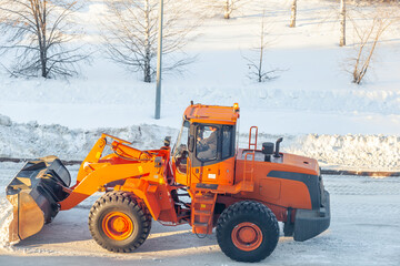 Obraz na płótnie Canvas Big orange tractor cleans up snow from the road and loads it into the truck. Cleaning and cleaning of roads in the city from snow in winter. Snow removal after snowfall and blizzards. 