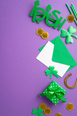 St Patrick's Day concept. Top view vertical photo of green envelope with letter irish party glasses small giftbox gold coins horseshoe straws and trefoils on isolated violet background