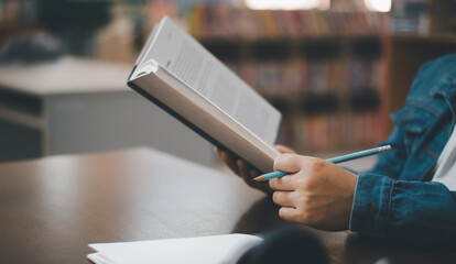 Closeup on female with book in the library in sunny day.Beautiful woman holding a book in hands while reading it on a bookshelf background, in a library.