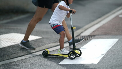 Fototapeta premium Child crossing street with scooter on zebra lines. Parent and kid on crosswalk in sunny day. Active small boy rides trandportation crosses sidewalk