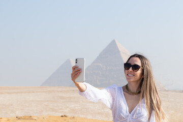 Young beautiful smiling tourist woman taking selfie picture of herself with the pyramid in blurred background