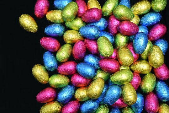 Pile or group of multi colored and different sizes of colourful foil wrapped chocolate easter eggs in pink, blue, yellow and lime green against a black background with copy space.