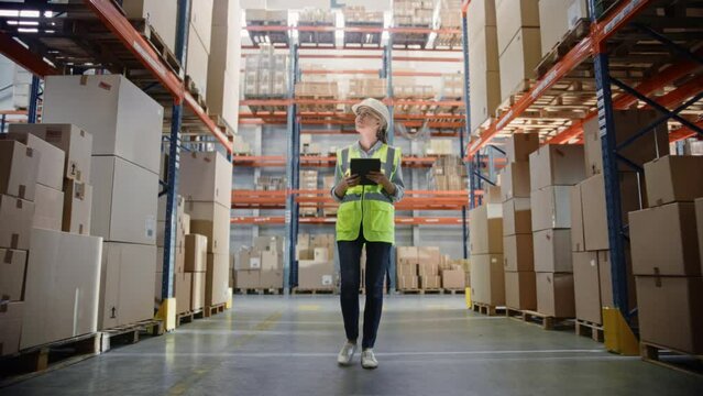 Professional Female Worker Wearing Hard Hat Uses Digital Tablet Checks Inventory Walks in the Retail Warehouse full of Shelves with Goods. Working in Logistics, Distribution Center. Following Shot