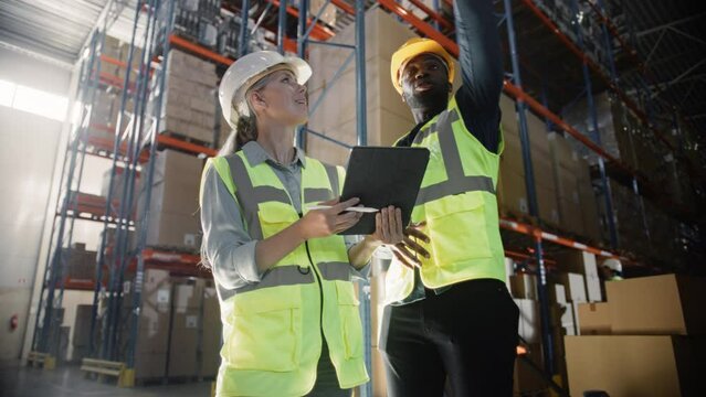 Black Male and Caucasian Female Supervisors Holding Digital Tablet Talk about Delivery Logistics in Retail Warehouse full of Shelves with Goods. Workers in Distribution and Delivery Center. Low Angle