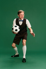Fototapeta na wymiar Portrait of man in stylish classical clothes, shorts and vest posing with football ball over green studio background. Concept of emotions, facial expression, lifestyle, retro fashion. Ad