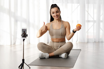 Fototapeta na wymiar Fitness blogger showing orange and thumb up, broadcasting from home