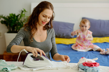 Smiling young woman ironng baby clothes when daughter playing in background