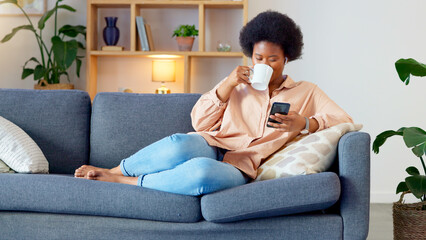 Laughing woman listening to music and sending a message with funny memes on her phone and drinking coffee while on the sofa. Happy black female browsing social media and surfing the internet online - obrazy, fototapety, plakaty