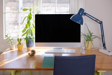 Pc computer monitor on the desk by the window with green plant decoration. Home office, freelancer workplace. Wooden table, lamp chair, work accessories, cozy remote work space arranged by home.