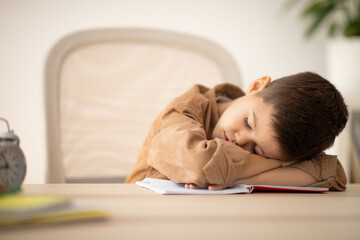 Tired small european boy lies at table sleeping, resting from lesson in school room interior