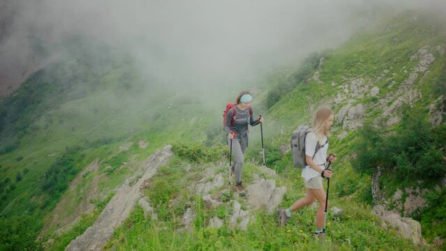 Two women with backpacks and trekking poles hiking in mountains amidst meadows 