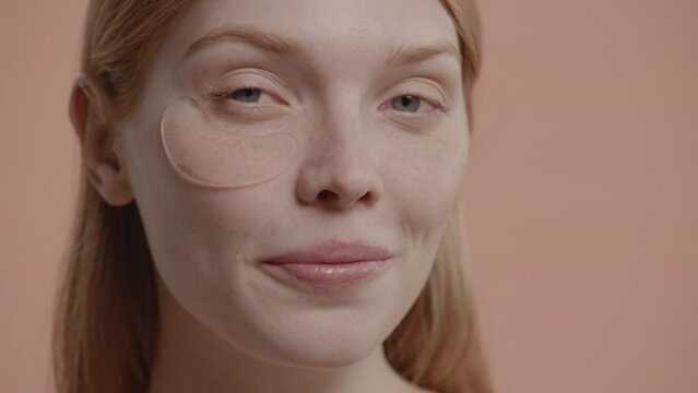 Portrait of pure natural beautiful 20s Caucasian female using eye patches against pastel colored background