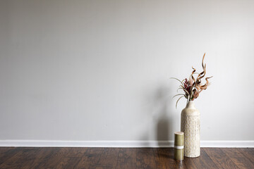 A tall clay vase filled with dried flower arrangements is situated on a laminated floor beside a candle in a white walled, empty room. It can serve as a backdrop for copy text.
