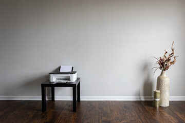 An interior with a white wall and laminated floor displays a small black table with an inkjet printer, and a tall clay vase filled with a dried flower arrangement next to a large candle.