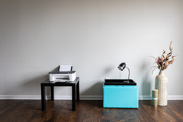 An interior of a home office featuring a small inkjet printer on a table, a stool with a lamp, and a tall clay vase filled with dried flowers on a laminated floor.