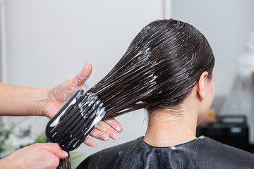 Naklejka premium Hairdresser applies a hair mask to straight black hair. Hair care at the beauty salon.