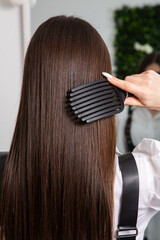 Young woman combing her long dark hair with a comb in a beauty salon. A straight healthy brunette hair that has undergone the hair straightening procedure.