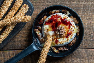 View of baked on frying pan camembert cheese, walnuts, honey, jam and bread sticks with sesame seeds on wooden table background, closeup, top view
