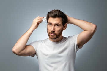 Morning preparation. Portrait of handsome middle aged man brushing his hair, looking at camera, grey background