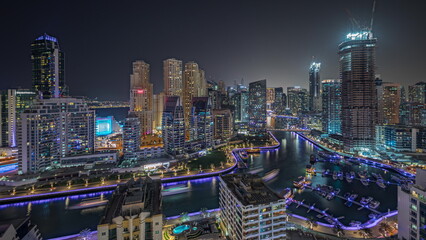 Panorama showing Dubai Marina with several boat and yachts parked in harbor and skyscrapers around canal aerial night .