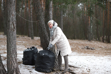 Portrait of happy beautiful elderly senior retired woman in age in forest or park at winter cold day, smiling, picking up garbage bag. Side view.The concept of environment protection and volunteering.