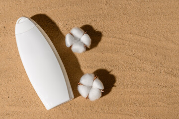 A white bottle of shampoo lies on the yellow sand. White cotton flowers.
