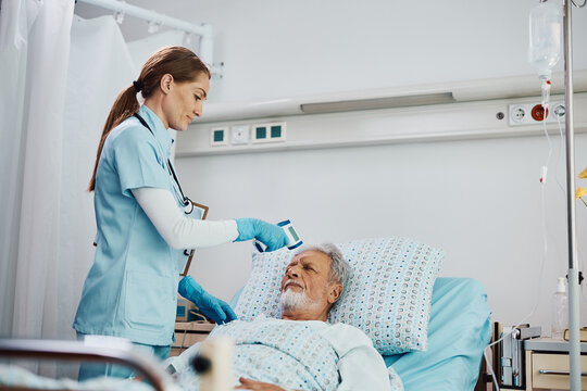 Smiling Nurse Measuring Senior's Patient Temperature In Hospital Ward.