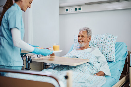 Happy senior patient talks to nurse who is bringing him food during his recovery in hospital.