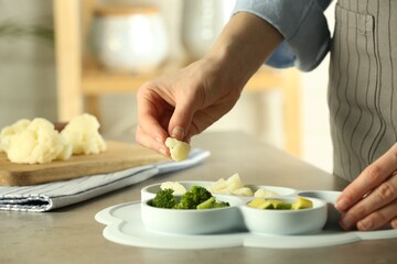 Baby food. Woman putting piece of cauliflower into section plate with different vegetables at grey textured table in kitchen, closeup