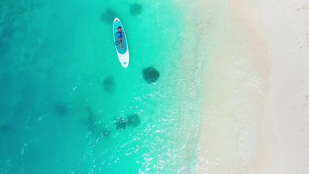 A little girl lying on a sup board in turquoise water with a white sand beach with stingray and sharks on Fulidhoo island in Maldives. Aerial top view