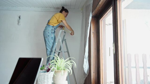A young caucasian woman is preparing a room for painting. A female fixing the film with masking tape while standing on a stepladder.