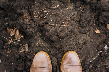 The worker is standing in two brown leather boots against the background of the earth, black soil, soil. Close-up photography, texture, farming, agriculture.