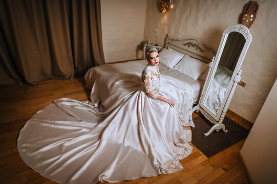 A beautiful young blonde model bride in a white long lace dress sits on a bed in a room in the morning. Wedding photography, portrait.