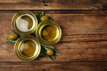 Bowls of cooking oil, ripe olives and green leaves on wooden table, flat lay. Space for text