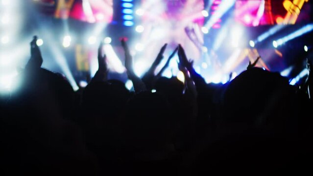 People clap their hands and dance at evening concert with light show.Fans raise their hands in bright colorful strobe lights on stage. Crowd is dancing, filming performance rock band, throwing party.