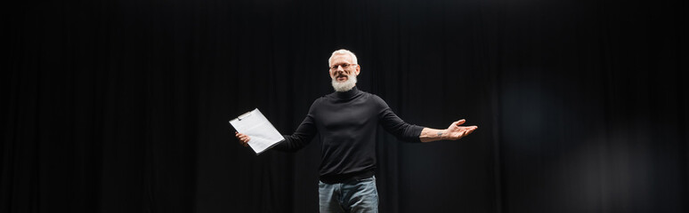 grey haired tattooed man in black turtleneck holding screenplay while rehearsing in theater, banner. Translation of tattoo: kanji, danger.