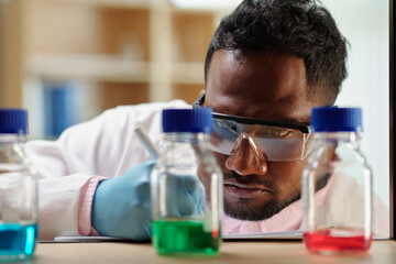 Serious scientist in protective glasses looking at bottles with reagents and taking notes in notebook