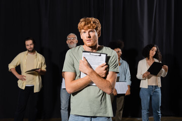 tensed redhead man holding scenario and looking at camera near multiethnic actors and producer on...