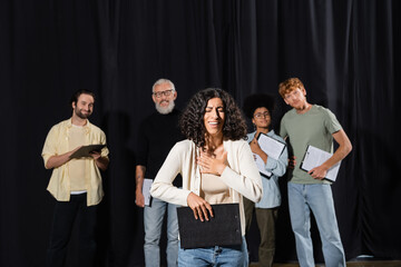 smiling multiracial woman holding clipboard and smiling near interracial actors and art director on...