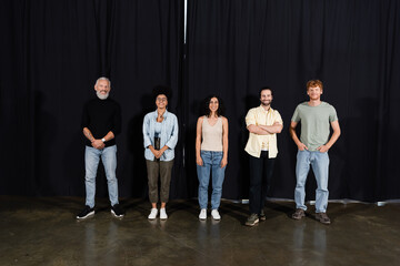 full length of happy art director with young multicultural actors standing on theater stage and looking at camera.