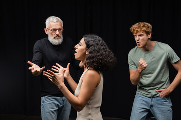 multicultural actress rehearsing near gesturing acting skills teacher and emotional redhead man in theater. - obrazy, fototapety, plakaty