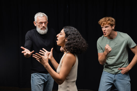 multicultural actress rehearsing near gesturing acting skills teacher and emotional redhead man in theater.